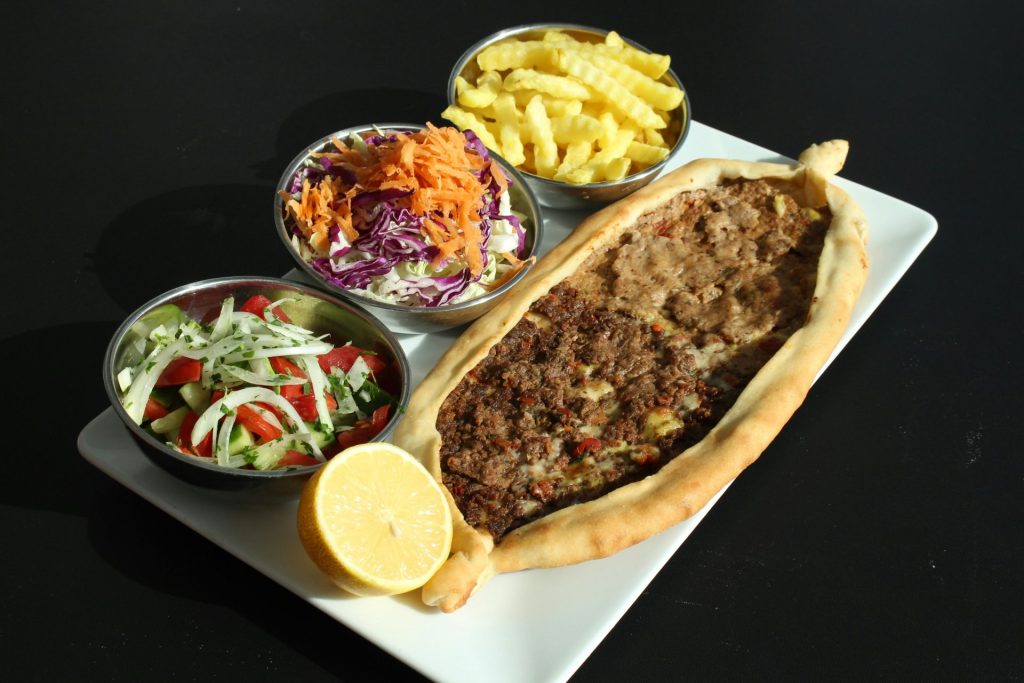 Ein weißer Teller mit einem fleischgefüllten Gebäckstück, daneben Schalen mit Salat und Pommes frites.